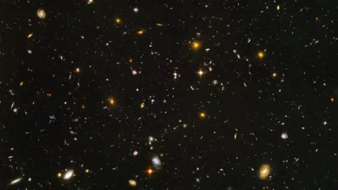 美国宇航局/欧洲航天局 这张图片展现了一片漆黑的夜空，上面点缀着成千上万个彩色的小点：每一个小点都是一个遥远的星系，而非一颗恒星。这些星系形状各异，色彩斑斓，从细小的蓝色光斑到较大的金色螺旋星系，再到模糊的红色斑点，均匀地散布在画面中。画面中没有前景或地平线，只有一片深邃无垠的宇宙景象，其中充满了各种各样的星系。